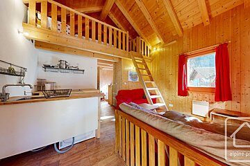 A cozy, rustic wooden cabin interior with a loft accessible by a ladder, a compact kitchenette, and a living area featuring a red sofa and red-curtained window.