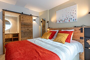 Cozy, modern rustic bedroom with a vibrant orange and yellow bed, wooden accents, a ski painting, and a barn door leading to a bathroom.