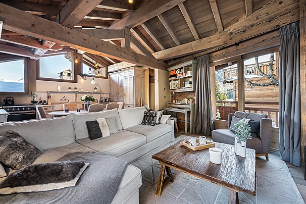 Cozy chalet living room with exposed wooden beams, a large sofa, open-plan dining/kitchen, and mountain views through large windows.