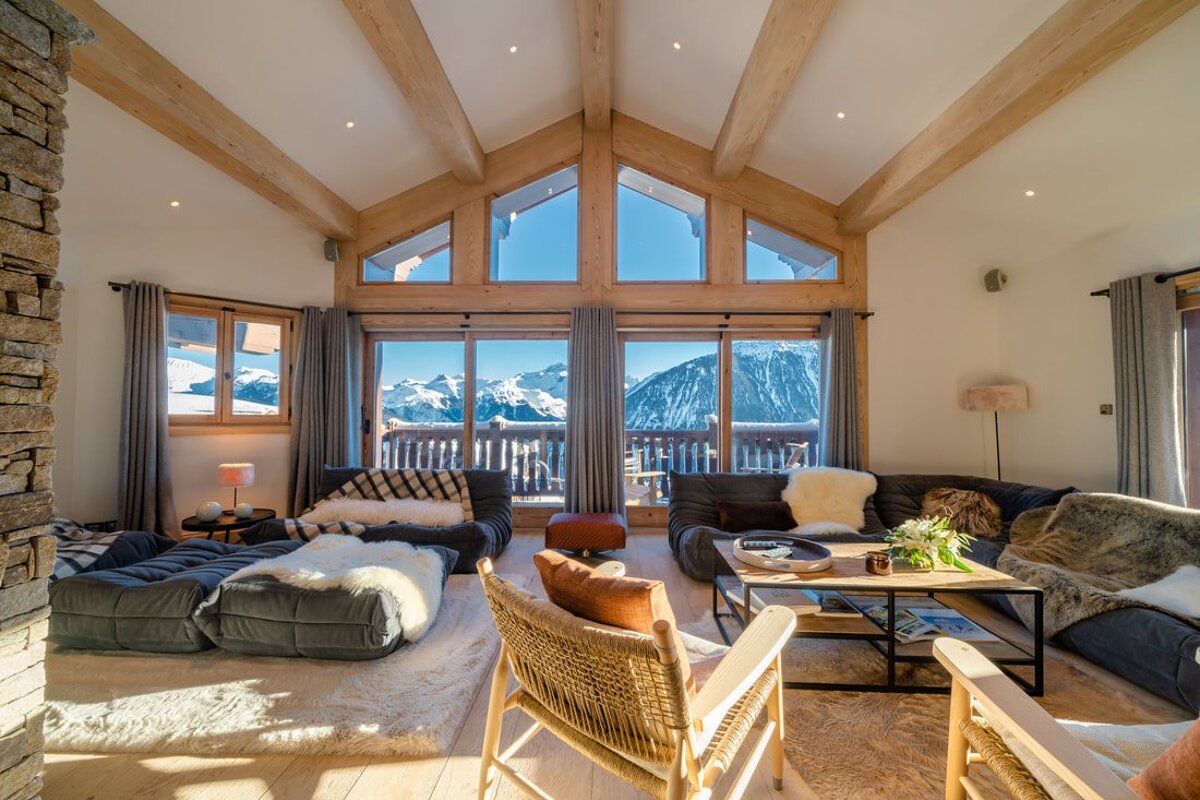 Cozy, rustic-modern living room with panoramic snowy mountain views through large windows, featuring wooden beams and plush seating.