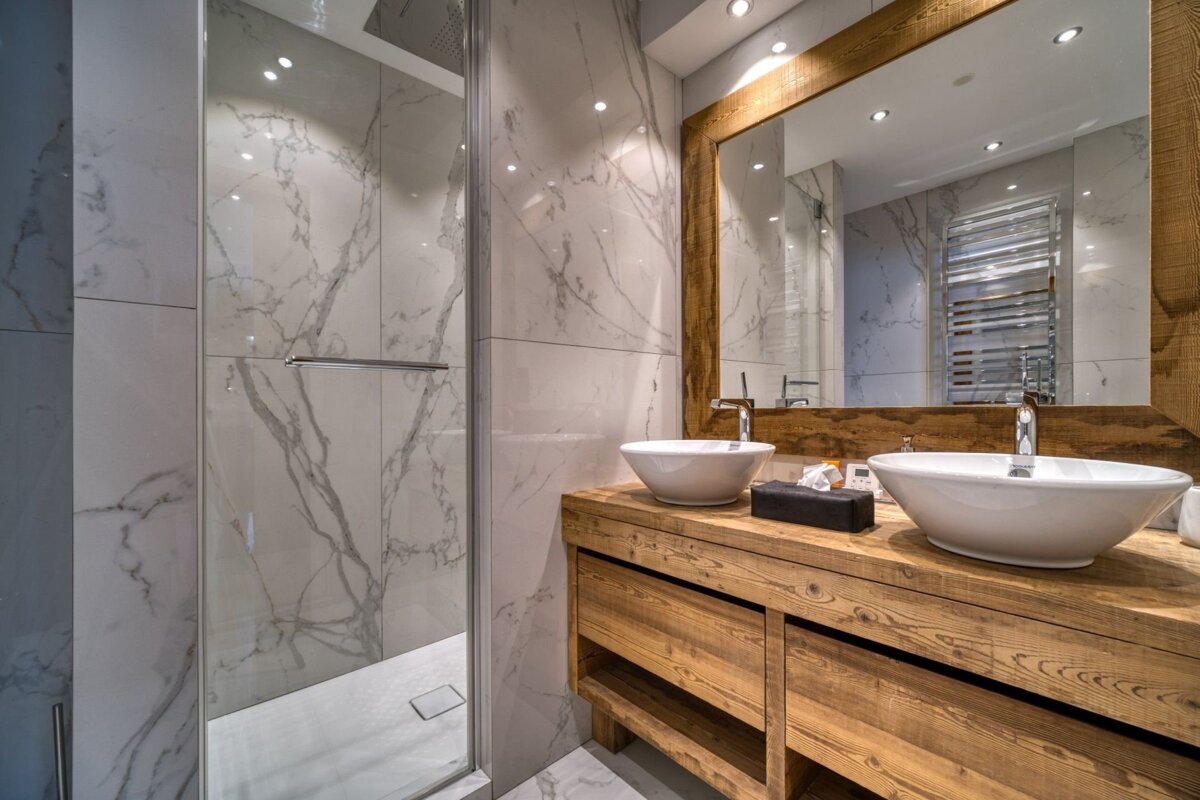 A modern bathroom with white marble walls, a wooden double vanity supporting two vessel sinks, a large wood-framed mirror, and a glass shower enclosure.