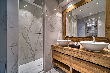 A modern bathroom with white marble walls, a wooden double vanity supporting two vessel sinks, a large wood-framed mirror, and a glass shower enclosure.