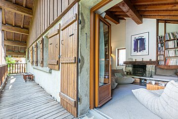 A wooden balcony with shutters opens into a cozy chalet living room featuring a fireplace, bookshelves, and beanbag chairs.