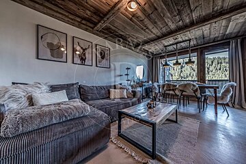 A warm, rustic chalet living/dining room with a grey sectional sofa, wood-paneled ceiling, fur-covered dining chairs, and a large window view of a forest.