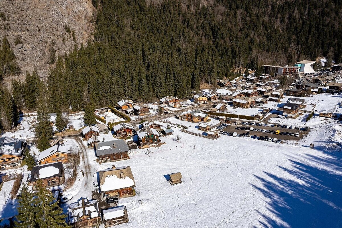 Aerial view of a snow-covered village with wooden chalets and a large parking lot, bordering a vast snowy expanse and a dense pine forest on a mountainside.
