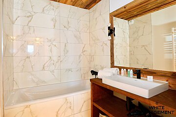 A modern bathroom featuring marble-look tiles, a built-in bathtub, and a wooden vanity with a sleek white sink and large mirror.