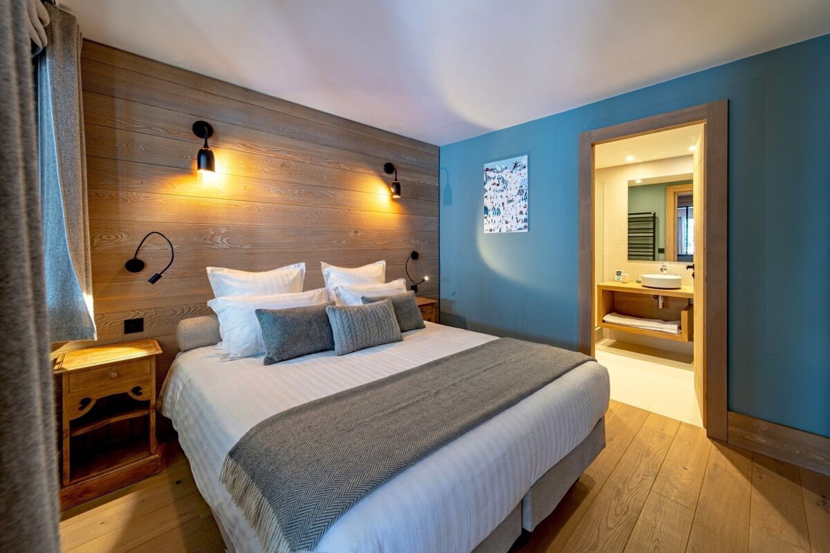 A cozy bedroom with a large bed, wooden headboard, blue accent wall, and ensuite bathroom. Warm lighting and wooden floors create a welcoming space.