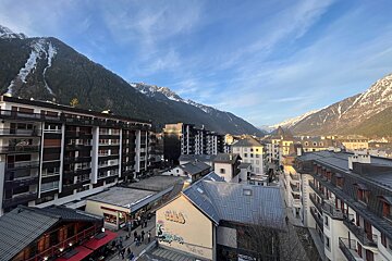 Elevated view of a bustling mountain town featuring varied architecture, nestled in a valley between snow-capped peaks under a bright blue sky.