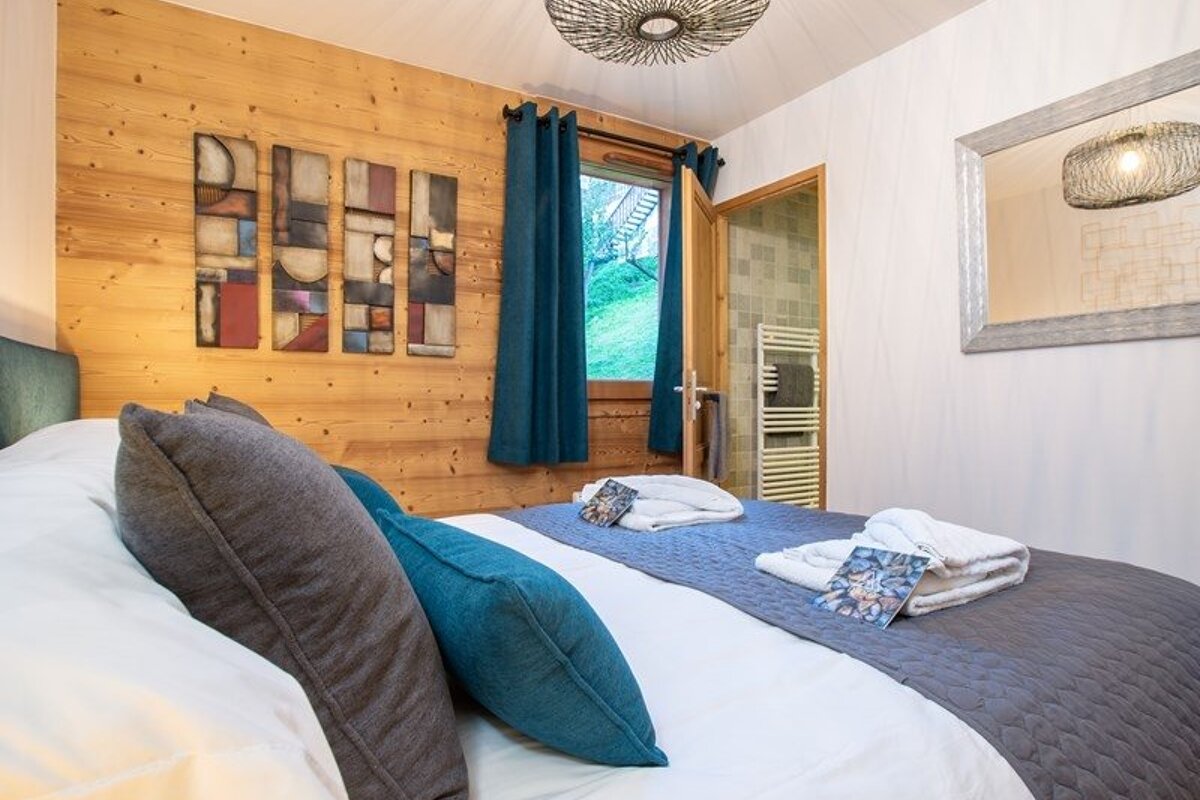 A rustic chalet bedroom featuring a wooden accent wall, a bed with blue and grey pillows, and teal curtains framing a window.