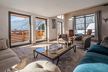Bright, modern living room with large windows and a balcony overlooking snowy mountains. Cozy, contemporary decor with stunning alpine views.