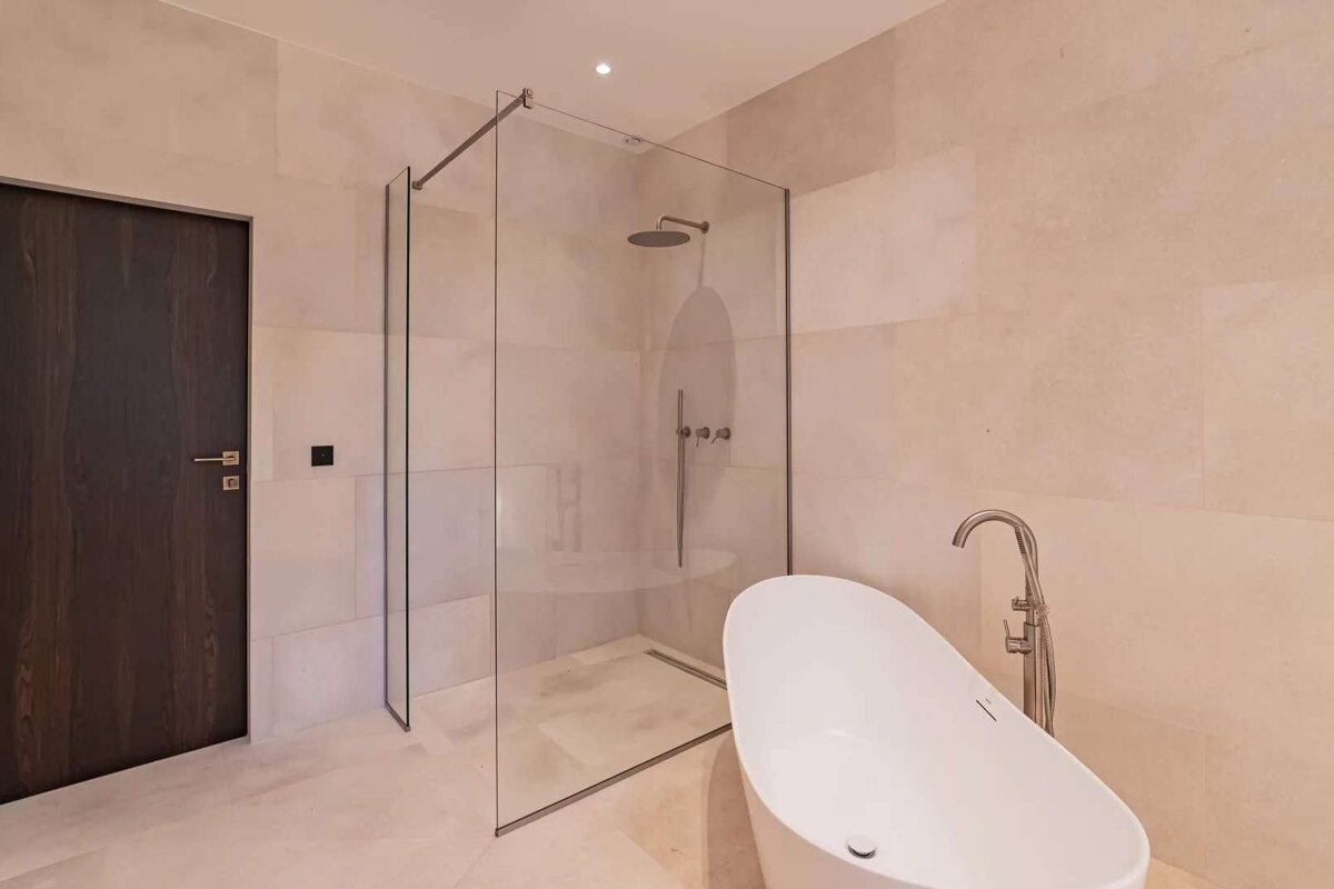 A sleek modern bathroom featuring a glass walk-in shower, a freestanding white bathtub, light beige tiled walls, and a dark wooden door.