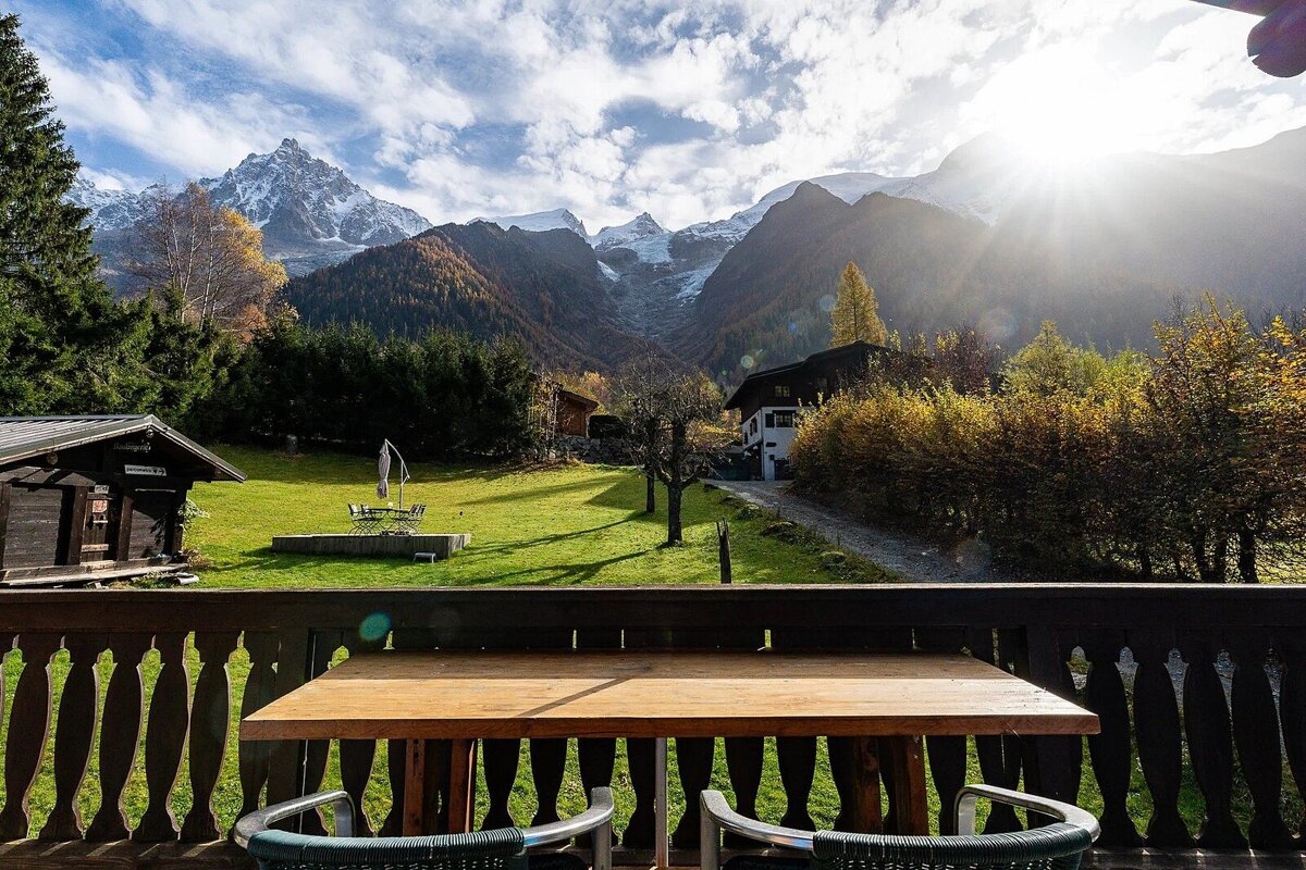 Sunny autumn view from a balcony overlooking a green lawn, chalets, and majestic sun-kissed, snow-capped mountains under a cloudy sky.