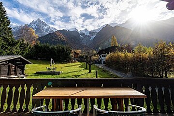 Sunny autumn view from a balcony overlooking a green lawn, chalets, and majestic sun-kissed, snow-capped mountains under a cloudy sky.