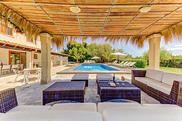 A sunny outdoor villa patio with wicker furniture under a thatched roof, overlooking a sparkling blue swimming pool surrounded by lush greenery.