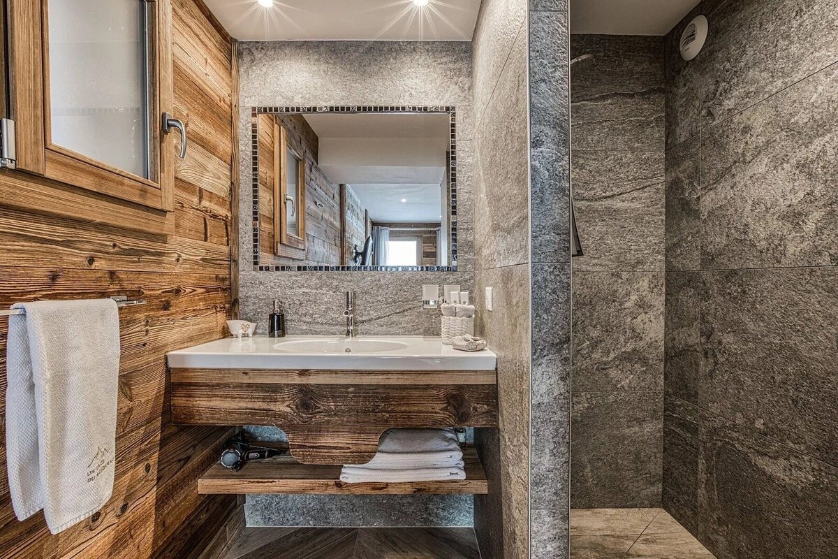 A cozy, modern bathroom with rich wooden walls and vanity, contrasting grey stone tiles, a sink, mirror, and shower area. Natural chalet style.