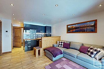 A modern open-plan kitchen and living room. Features dark blue kitchen, grey L-shaped sofa with purple accents, wood flooring, and mountain art.