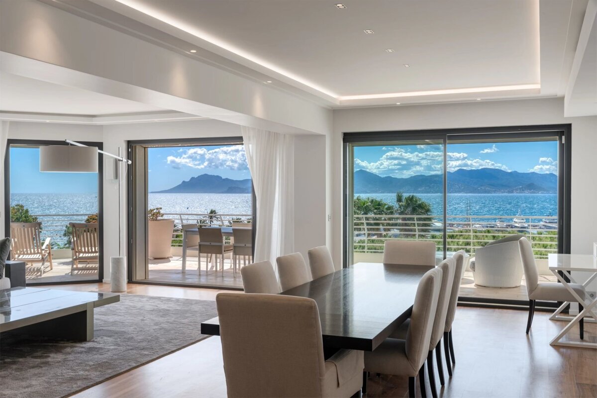 Modern dining room and living area with a dark table, plush chairs, and floor-to-ceiling windows offering a panoramic ocean and mountain view.