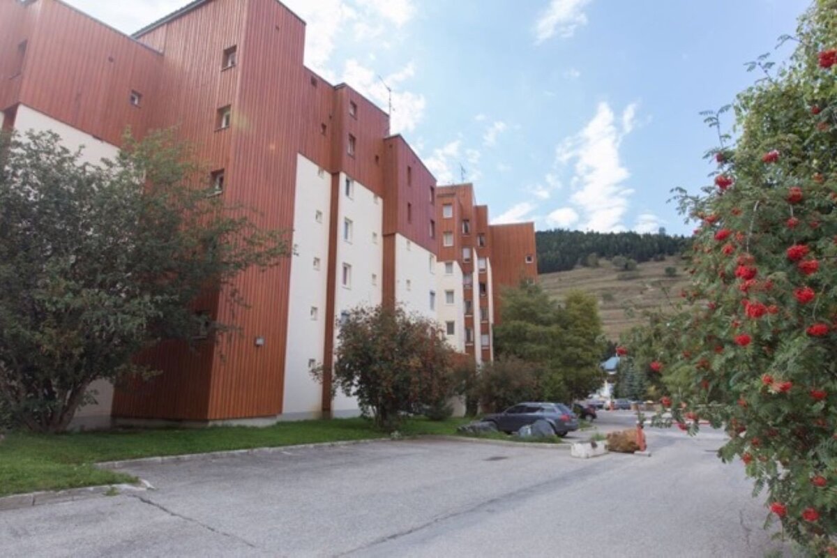 A large brown and white building stands beside a parking lot. Trees with red berries frame the right, while a forested mountain rises under a blue sky.
