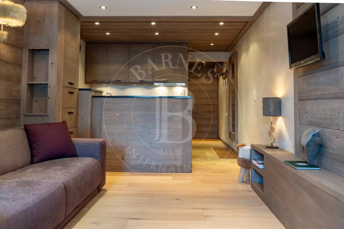 A cozy, modern chalet-style room with extensive light wood paneling, light floors, a sofa, and a compact kitchen. Warm lighting, a TV, and neutral tones.