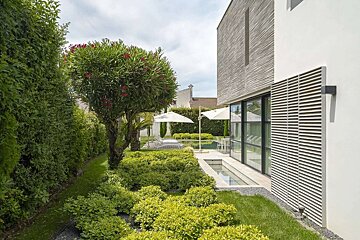 A modern house with textured walls overlooks a lush green garden featuring a flowering tree, patio with umbrellas, and a sleek water feature.