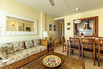 A bright living-dining room featuring a wicker sofa, floral cushions, wooden dining set, and a kitchen pass-through. Coastal decor adorns the coffee table.