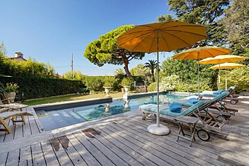 A sunny swimming pool area with wooden decking, blue lounge chairs, yellow umbrellas, and green trees.