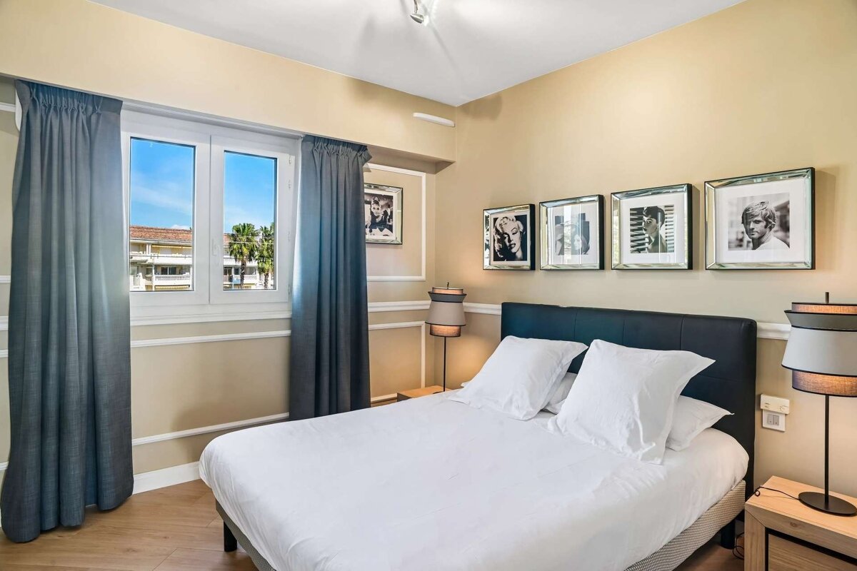 A cozy bedroom with a neatly made bed, window with blue curtains overlooking palm trees, and framed black-and-white celebrity photos on beige walls.