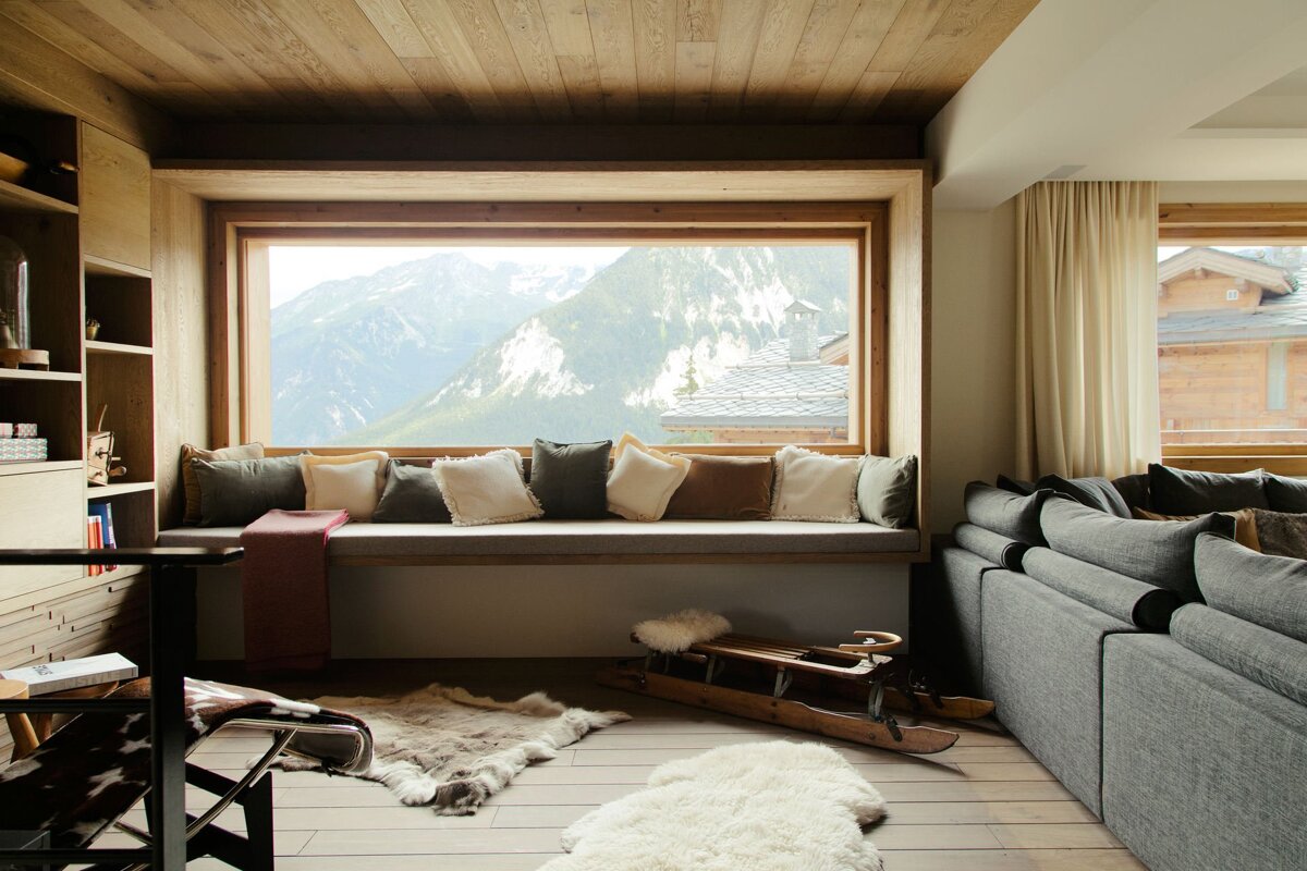 A cozy, rustic chalet living room with a large window seat overlooking stunning snowy mountains, featuring wooden accents, plush cushions, and fur rugs.