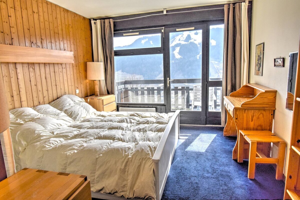Cozy wood-paneled bedroom with a large bed, blue carpet, and balcony offering stunning snowy mountain views. Features a wooden desk.