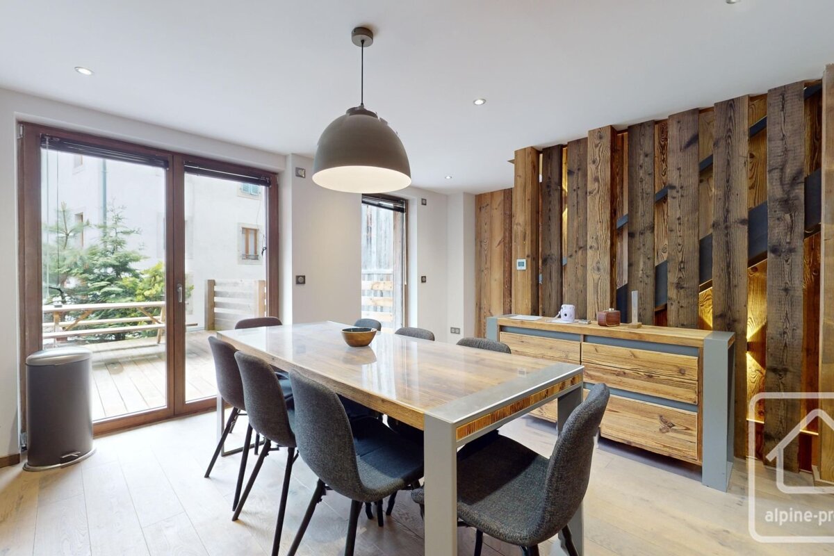 A modern chalet-style dining room features a large wooden table with grey chairs, a unique wooden accent wall with lighting, and glass doors to a balcony.