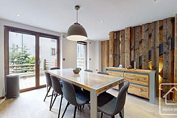 A modern chalet-style dining room features a large wooden table with grey chairs, a unique wooden accent wall with lighting, and glass doors to a balcony.