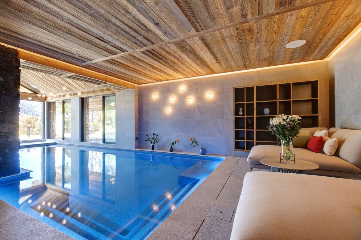 A modern indoor swimming pool with a warm wooden ceiling, large windows, elegant lighting, and a cozy lounge area with shelving.