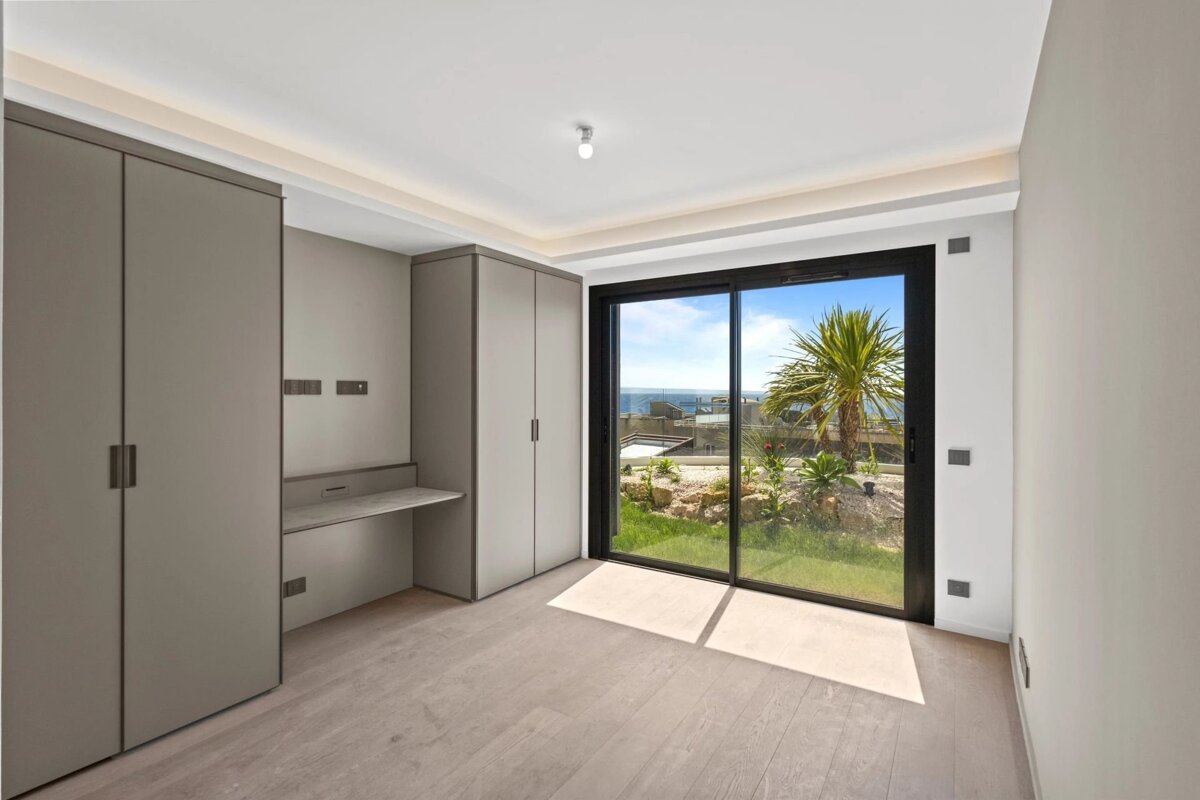Modern empty room with grey wardrobes, a built-in desk, and sliding glass doors opening to a sunny garden with a palm tree.