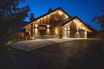 A stunning, illuminated wooden and stone mountain chalet at dusk. Warm lights highlight its features, with a large stone and asphalt driveway.