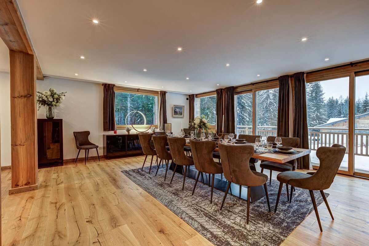 A modern, spacious dining room features a long table with brown velvet chairs, set against large windows offering a snowy forest view. Wooden floors and warm lighting create a cozy ambiance.