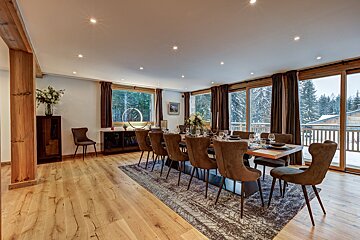 A modern, spacious dining room features a long table with brown velvet chairs, set against large windows offering a snowy forest view. Wooden floors and warm lighting create a cozy ambiance.