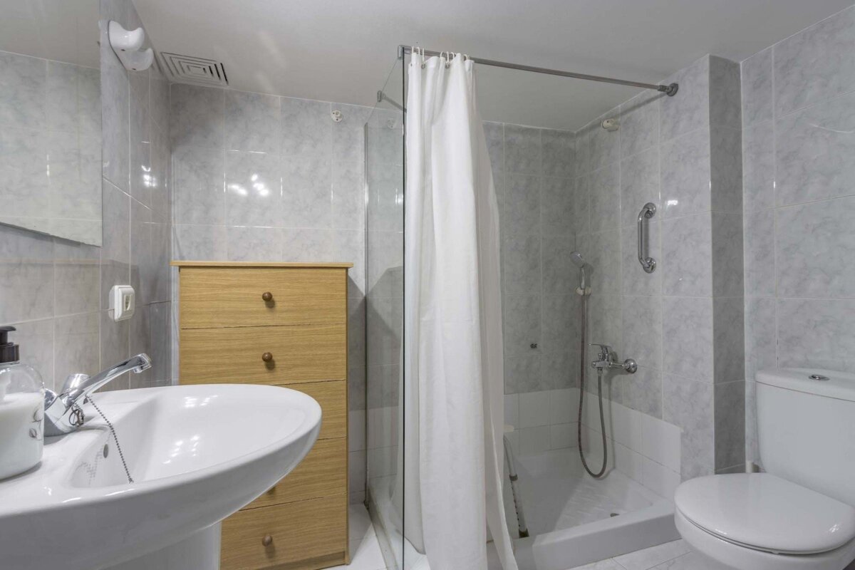 A clean, grey-tiled bathroom featuring a white pedestal sink, a wooden drawer chest, a shower with a white curtain, and a toilet.