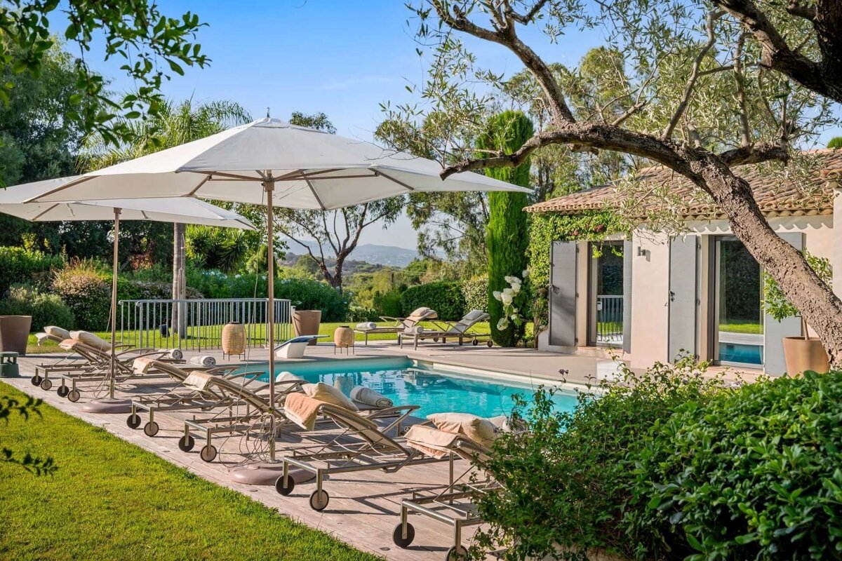 Sunny outdoor pool with lounge chairs, white umbrellas, and a beautiful villa nestled in a green garden with distant hills.