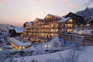 Luxurious wooden chalets in a snow-covered mountain resort glow warmly at dusk, nestled amidst mountains and frosted trees.
