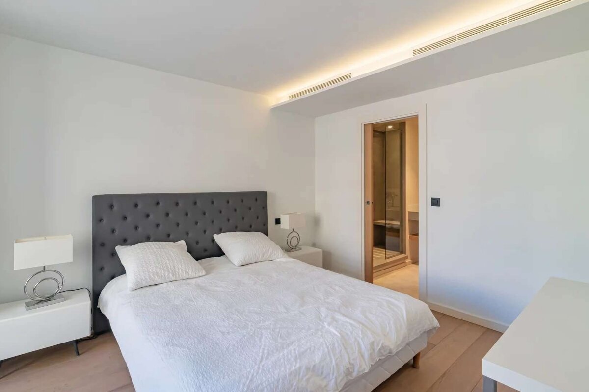 A bright, modern bedroom with white walls, a gray tufted bed, and light wood floors. An en-suite bathroom with a glass shower is visible.