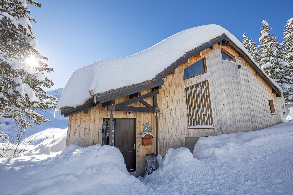 A cozy wooden cabin blanketed in deep snow, surrounded by snow-covered pines under a bright blue sky with a shining sun.