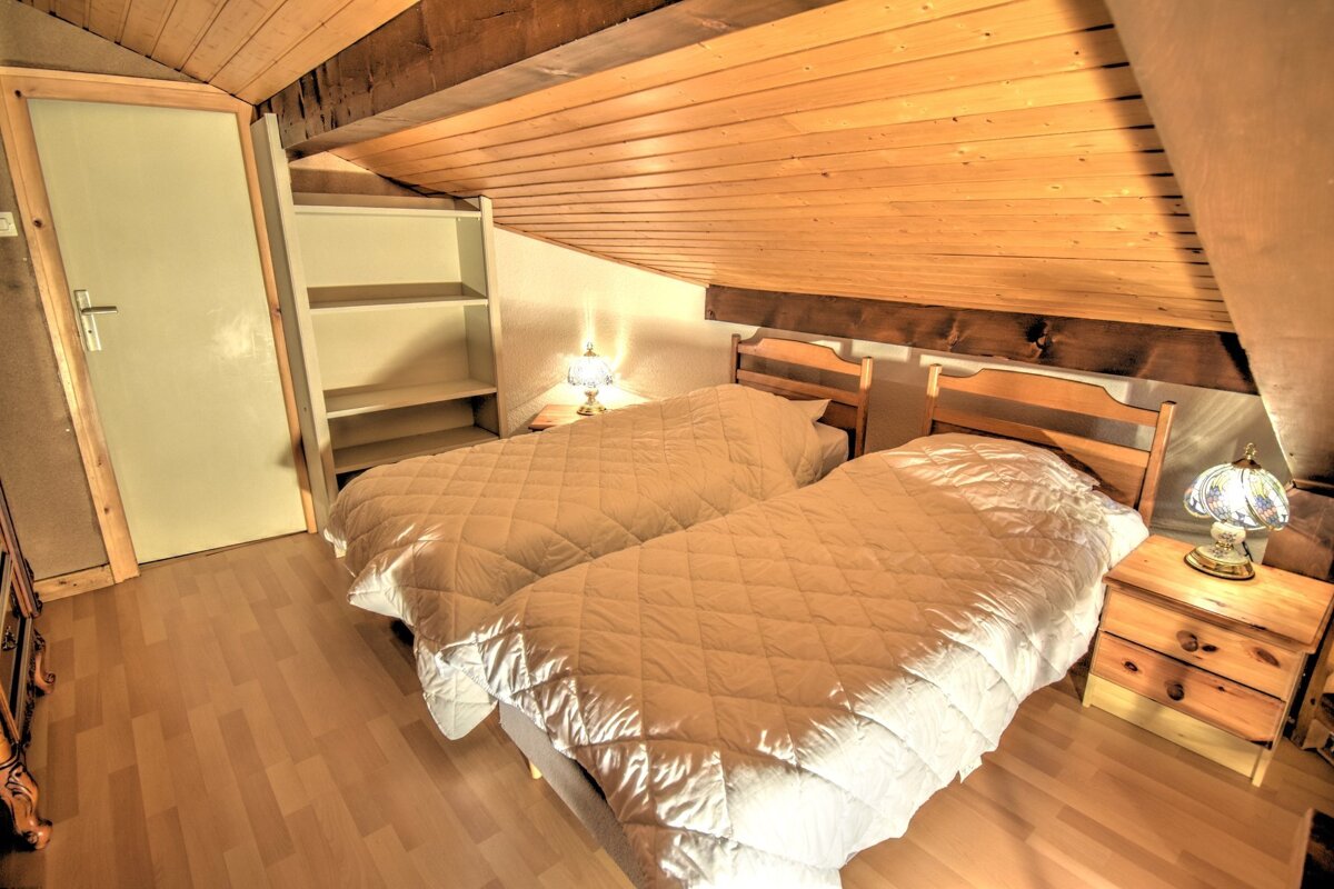Cozy attic bedroom featuring two twin beds with white duvets, slanted wooden paneled ceilings, and warm bedside lighting.