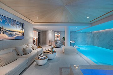A modern, luxurious indoor spa and lounge featuring a white sofa, an illuminated pool, a large TV, and soft blue accent lighting.