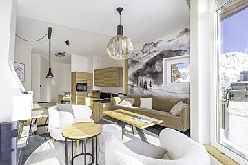 A bright, modern chalet living room featuring white armchairs, a mountain wall mural, and large windows showcasing a snowy alpine view.