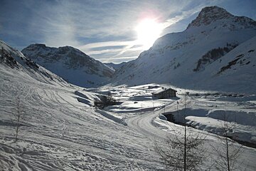 A sunlit, snow-covered mountain valley with a wooden building, winding road, and ski tracks under a blue sky with thin clouds.