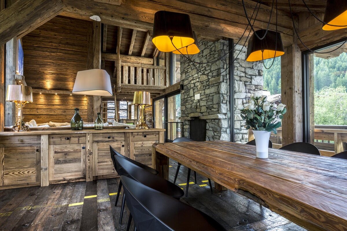 A rustic chalet dining room with a long live-edge wooden table, modern lighting, stone fireplace, and large windows offering a forest view.