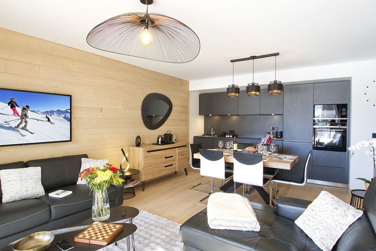 A modern open-plan living, dining, and kitchen area with a wood-paneled wall. Features black sofas, a TV with a ski scene, and a dark gray kitchen.
