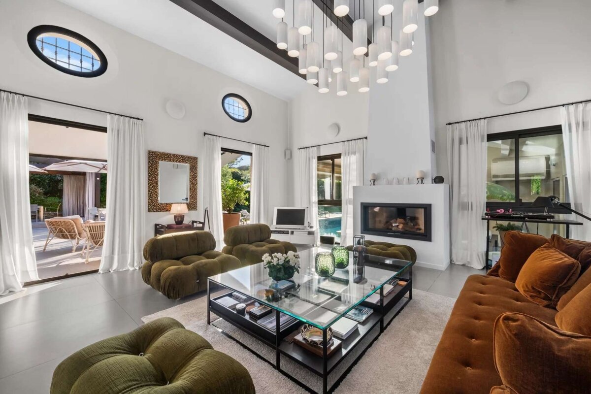 Spacious, luxurious living room with high ceilings, a modern chandelier, fireplace, plush velvet seating, and large windows leading to a sunny patio and pool.