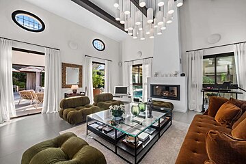 Spacious, luxurious living room with high ceilings, a modern chandelier, fireplace, plush velvet seating, and large windows leading to a sunny patio and pool.