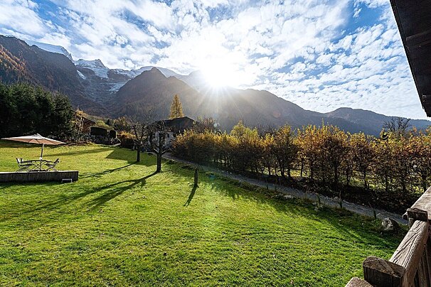 Sun-drenched mountain valley with snow-capped peaks and a vibrant green lawn. An outdoor dining set sits under an umbrella, framed by autumn trees.